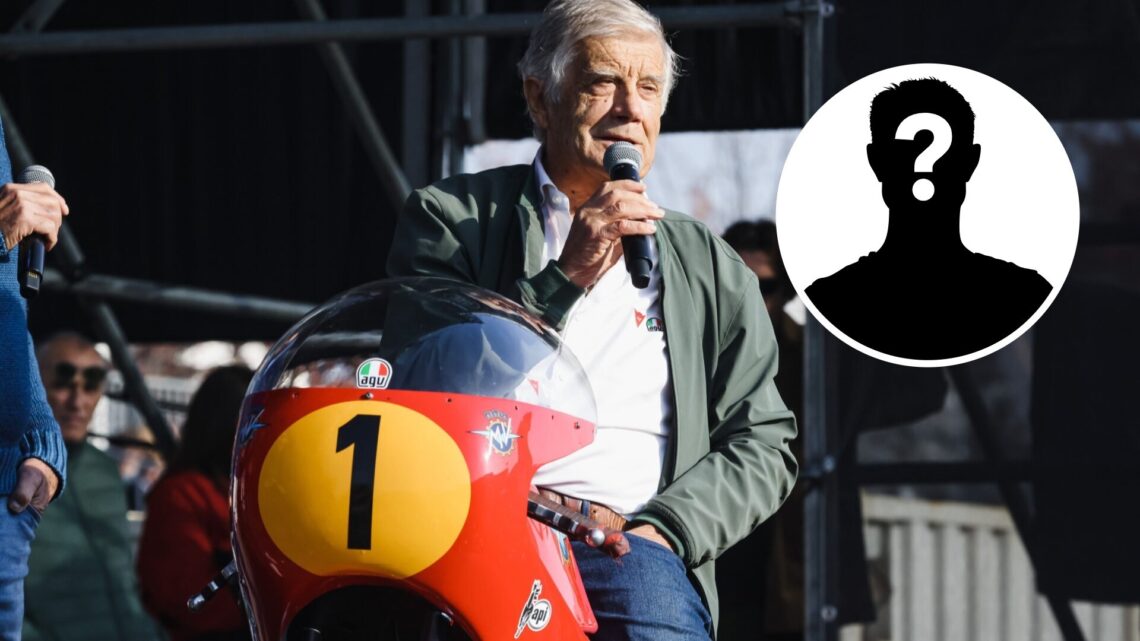Giacomo Agostini speaking into a microphone at a motorcycle event in Milan.
