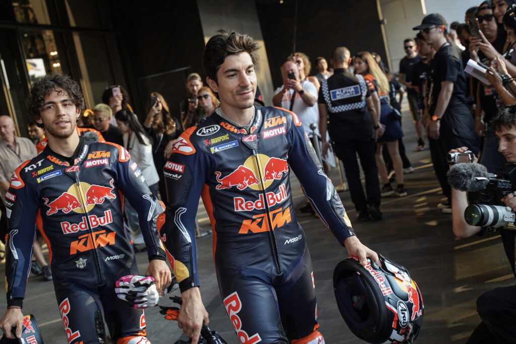 Maverick Vinales and Enea Bastianini walk towards the camera, carrying their helmets at the 2025 MotoGP season launch event as crowds take photos